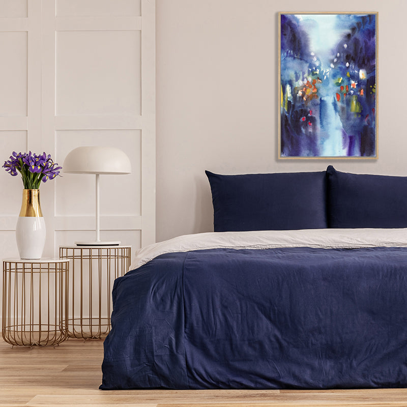 Indigo blue art print, capturing the glow of street lights on a rainy night in the city, above a bed with navy covers. 
