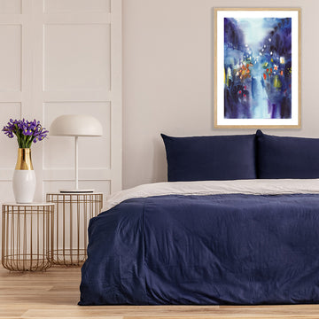 Framed art print featuring indigo blue, capturing the glow of street lights on a rainy night, above a bed with navy covers.
