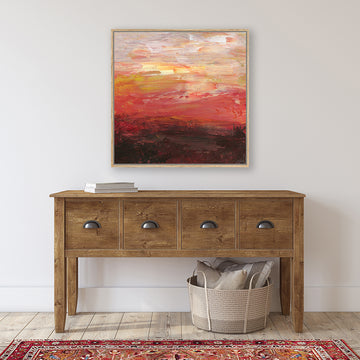 A framed abstract canvas art print featuring a fiery brick-red sunset above a deep mahogany outback landscape, displayed on a wall above a wooden console table with a red Persian rug in front.